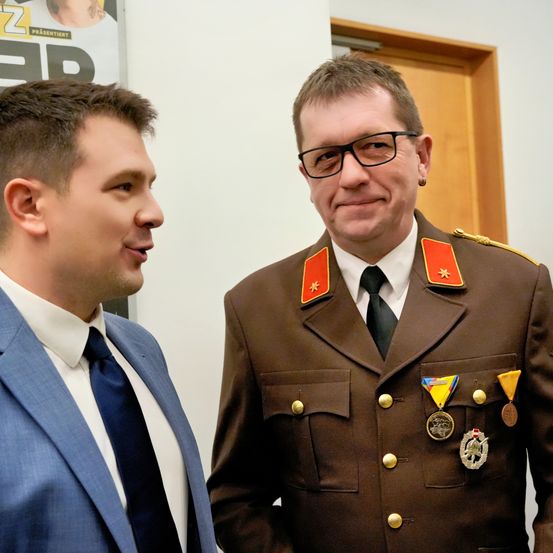 Zwei Männer stehen nebeneinander, einer in einem Anzug und der andere in einer Militäruniform mit Medaillen. Sie lächeln und scheinen zu sprechen. Hinter ihnen ist ein Poster mit einer Tätowierung darauf.