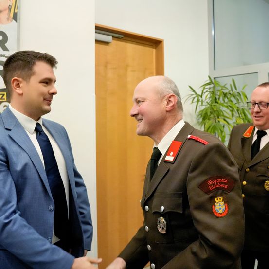 Zwei Männer in Uniformen und ein Mann im Anzug schütteln Hände. Sie lächeln und scheinen sich zu unterhalten. Hinter ihnen steht eine Holztür und eine Topfpflanze.