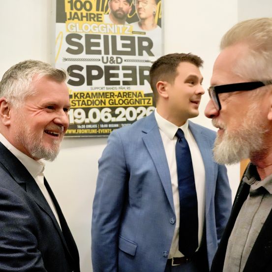 Drei Männer in Anzügen und Krawatten lächeln und unterhalten sich vor einem Plakat, das ein Konzert von Seiler und Speer bewirbt.