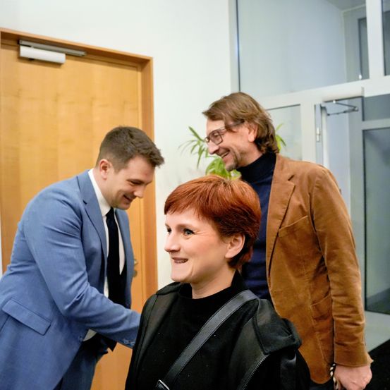 Drei Personen stehen in einem Büro; ein Mann in einem blauen Anzug lächelt, eine Frau mit roten Haaren lächelt und ein Mann in einem braunen Mantel lacht. Hinter ihnen ist eine Holztür und eine Pflanze zu sehen.