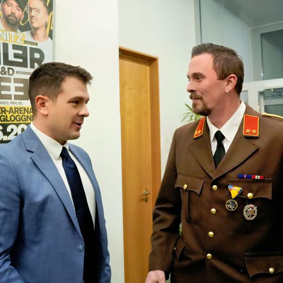 Zwei Männer stehen sich in einem Raum gegenüber. Einer trägt eine braune Militäruniform mit Medaillen, der andere einen blauen Anzug. Hinter ihnen befindet sich ein Poster mit verschwommenem Text und eine Holztür.