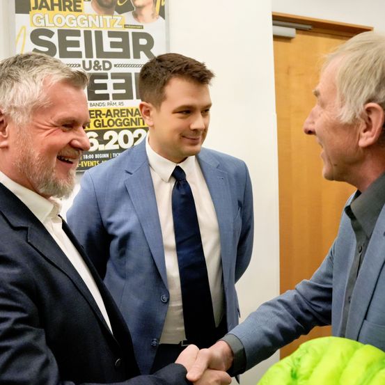 Drei Männer in Anzügen stehen eng beieinander, zwei schütteln sich die Hände und lächeln. Ein Poster hinter ihnen trägt den Text 'Jahre Gloggnitzer' und ein Datum, mit einem Bild eines Mannes.