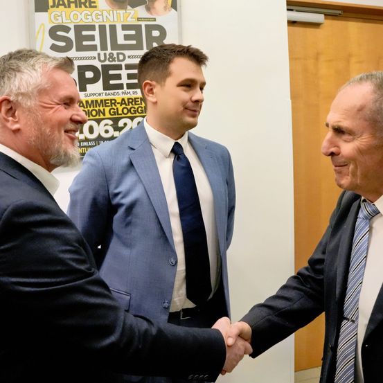 Drei Männer in Anzügen schütteln sich die Hände in einem Raum, mit einem Poster im Hintergrund.