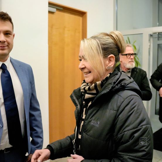 Ein Mann und eine Frau lächeln und schauen sich an. Sie tragen beide Jacken und Schals. Der Mann trägt Anzug und Krawatte. Hinter ihnen ist eine Tür und ein Mann mit Brille.