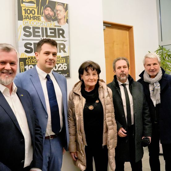 Fünf Erwachsene, drei Männer und zwei Frauen, stehen nebeneinander und lächeln. Sie sind formell gekleidet. Hinter ihnen öffnet sich eine Tür zu einem Korridor. An der Wand ist ein Poster mit dem Text '100 Jahre Gloggnitzer Seiler' und Bildern von zwei Männern angebracht.