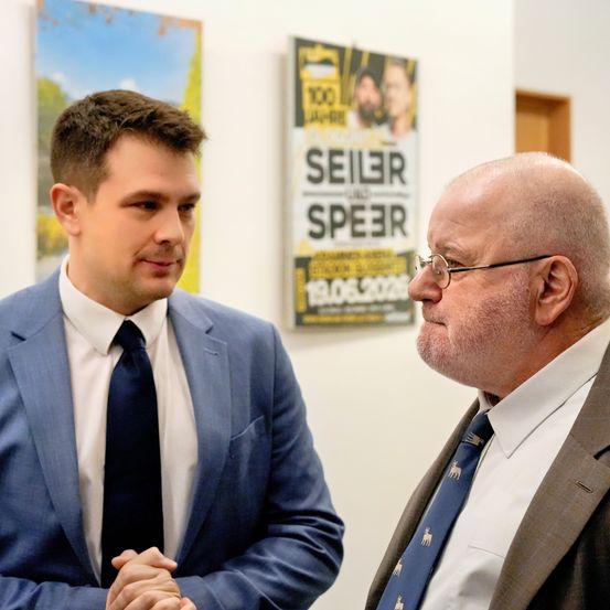 Zwei Männer in Anzügen unterhalten sich vor einem Plakat mit den Worten 'Seiler und Speer' und dem Datum '19.06.2020'.