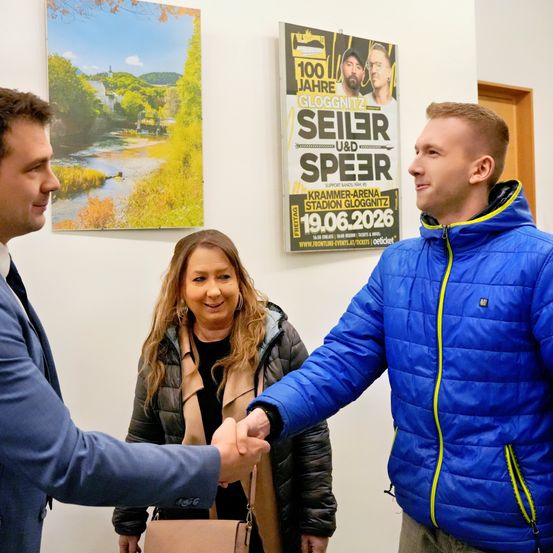 Drei Menschen sind in einem Raum, zwei Männer und eine Frau. Sie schütteln sich die Hände. Die Frau lächelt. Hinter ihnen ist ein Poster.