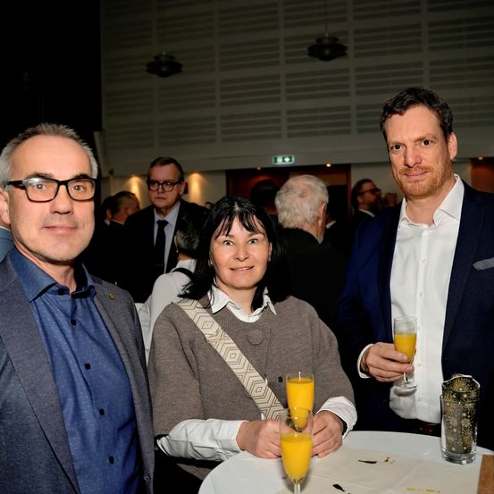 Drei Personen stehen bei einem Tisch mit Orangensaft in Gläsern und lächeln. Ein Mann links, eine Frau in der Mitte und ein Mann rechts. Hinter ihnen ein verschwommener Hintergrund von Menschen.