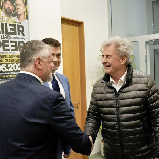 Drei Männer in Anzügen und Krawatten, einer in einer schwarzen Daunenjacke, schütteln sich die Hände in einem Büro, mit einem Plakat dahinter.