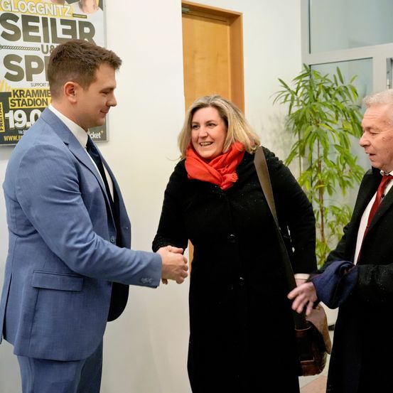 Drei Menschen stehen drinnen, wobei zwei Männer sich die Hände schütteln und eine Frau in die Kamera lächelt. Der Mann auf der linken Seite trägt einen blauen Anzug, während die anderen beiden in formeller Kleidung sind. Ein Plakat mit dem Wort 'Seiler' ist hinter ihnen sichtbar.