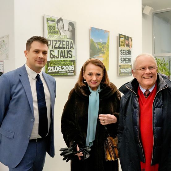 Drei Personen stehen nebeneinander vor einer Wand mit Werbeplakaten und lächeln für ein Foto.