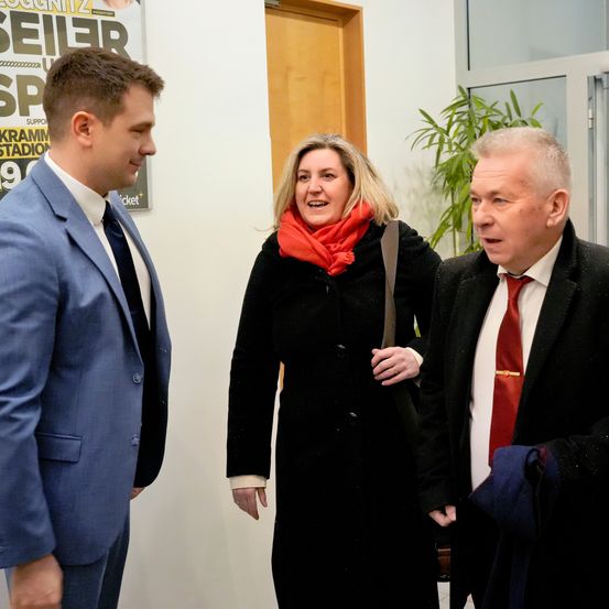 Drei formell gekleidete Personen, möglicherweise in einer Besprechung, wobei eine Frau lächelt und ein Mann seinen Mantel hält. Ein Plakat an der Wand lautet 'Sei1er'.