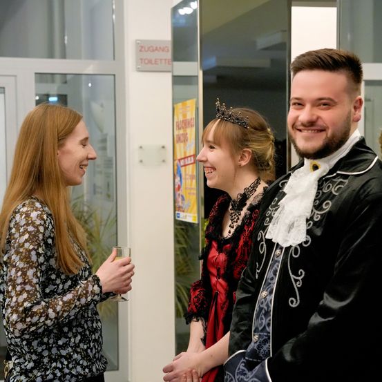 Drei Personen stehen nebeneinander. Die Frau auf der linken Seite lächelt und hält ein Glas. Die Frau in der Mitte trägt eine Krone. Der Mann auf der rechten Seite trägt ein Kostüm. Hinter ihnen ist eine Glastür mit einem Schild, das 'Zugang Toilette' steht.