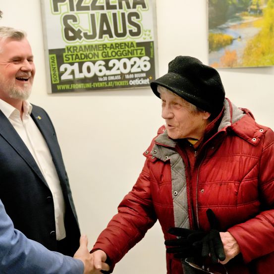 Zwei Männer und eine ältere Frau stehen eng beieinander. Der Mann in einem schwarzen Anzug und einem weißen Hemd lächelt. Die Frau trägt eine rote Jacke und einen schwarzen Hut. Sie schütteln sich die Hände. Hinter ihnen ist ein Plakat für eine Pizza- und Jaus-Veranstaltung am 21. Juni 2026 an der Wand angebracht.