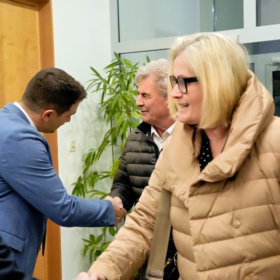 Drei Personen sind drinnen. Ein Mann im blauen Anzug schüttelt die Hand einer Frau im braunen Mantel. Hinter ihnen steht ein älterer Mann mit weißem Haar, der einen schwarzen Mantel trägt. Im Hintergrund befindet sich eine Pflanze und eine Tür.