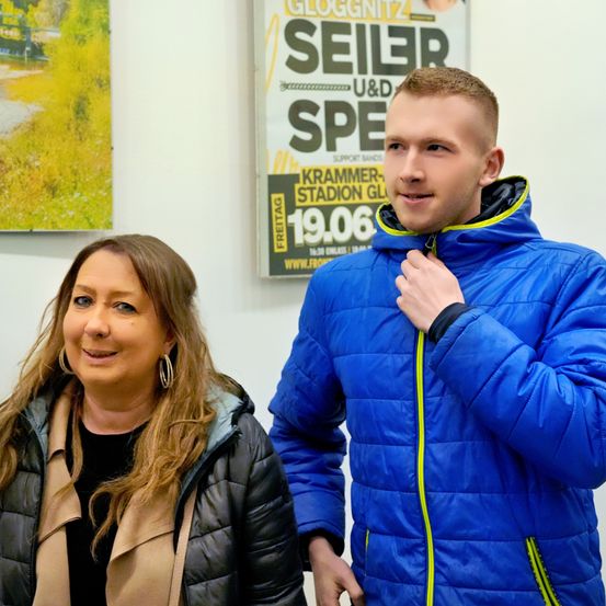 Ein Mann und eine Frau stehen lächelnd in einem Raum. Der Mann trägt eine blaue Jacke und die Frau eine schwarze Jacke. Hinter ihnen ist ein Poster mit dem Wort Seiler.