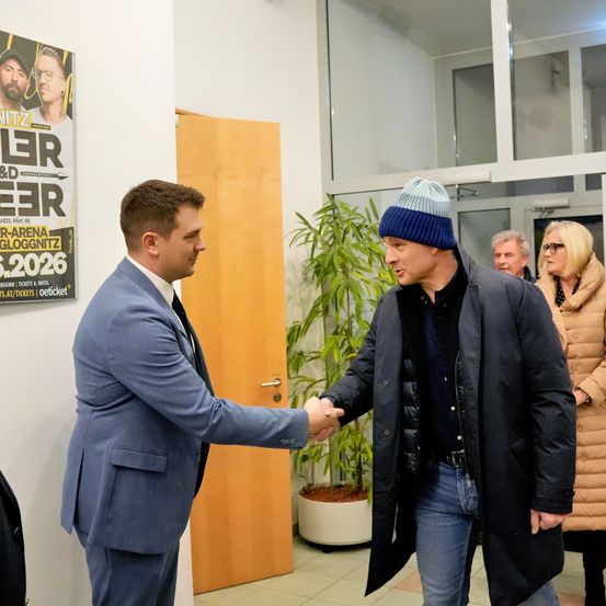 Zwei Männer schütteln sich die Hände in einer Lobby; einer in Anzug und der andere in einer Mütze. Hinter ihnen wirbt ein Plakat für eine Veranstaltung.