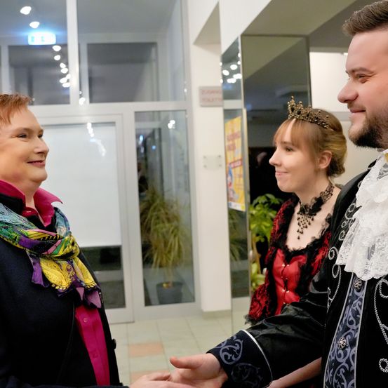 Eine Frau in einem bunten Schal begrüßt einen Mann in einem aufwendigen Kostüm, während eine Frau in einem roten Kleid in einem Raum mit Glastüren und einem Spiegel zusieht.