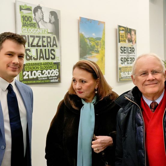 Drei Personen stehen vor einer Wand mit Plakaten, eines mit '100 Jahre Gloggnitz', ein weiteres mit 'PIZZERA & JAUS' und ein drittes mit '100 Jahre Seiler und Speer'.