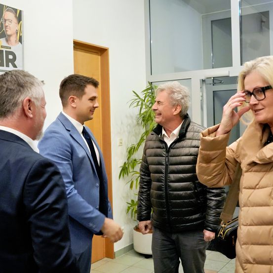 Eine Gruppe von Menschen steht lächelnd in einem Raum. Eine Person trägt eine Brille und eine braune Jacke. Ein Poster ist an der Wand angebracht.