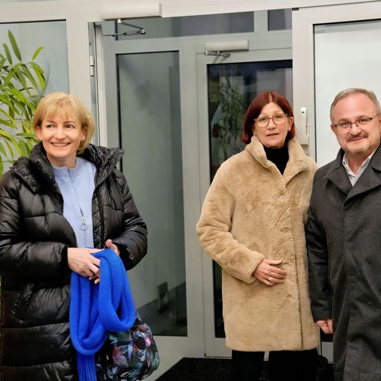 Drei Personen, zwei Frauen und ein Mann, stehen vor einem Gebäude. Die Frau auf der linken Seite hält einen blauen Schal, und die Frau in der Mitte trägt eine Brille. Der Mann trägt eine Brille und einen Ring.