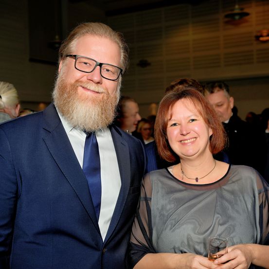 Ein Mann und eine Frau in formeller Kleidung lächeln und posieren für ein Foto bei einer Veranstaltung. Der Mann hat einen Bart und Brille, während die Frau ein Glas hält. Hinter ihnen sind verschwommene Menschen im Hintergrund zu sehen.