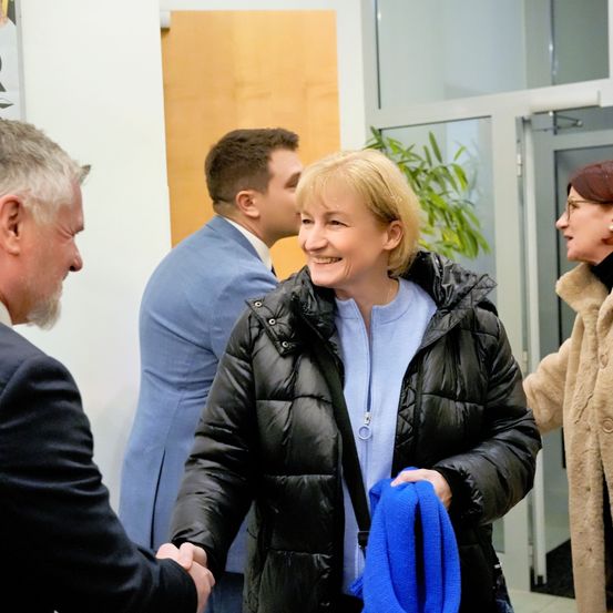 Eine Frau in einer schwarzen Steppjacke hält einen blauen Schal und lächelt, während sie einem Mann im Anzug die Hand schüttelt. Hinter ihr unterhalten sich zwei weitere Personen. Im Hintergrund sind eine Wand und Glastüren zu sehen.