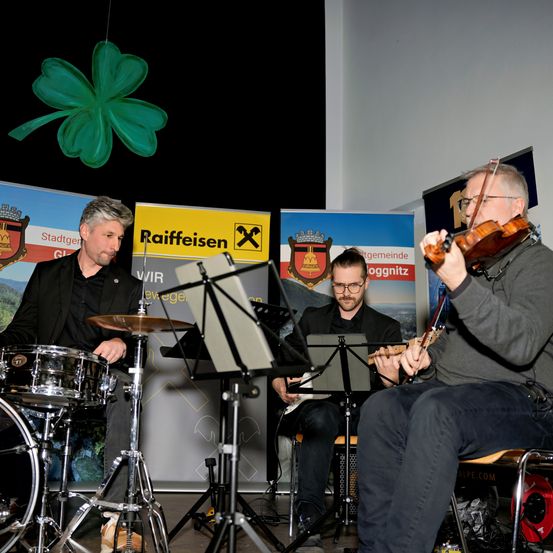 Drei Männer spielen Instrumente auf einer Bühne, einer mit einer Geige, einer mit einer Gitarre und einer mit einem Schlagzeug. Sie treten vor einer weißen Wand mit Bannern auf.