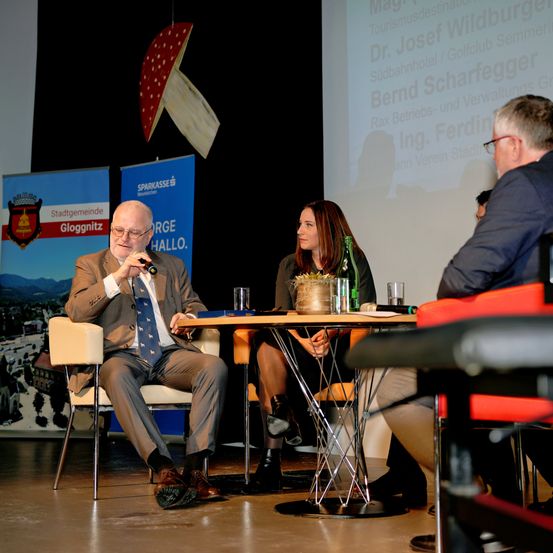 Ein Mann spricht in ein Mikrofon an einem runden Tisch mit zwei Frauen. Hinter ihnen ist eine Projektionsfläche mit Text.