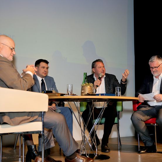 Vier Männer in Anzügen sitzen um einen Tisch herum. Einer spricht in ein Mikrofon. Die anderen hören aufmerksam zu. Der Mann rechts hält ein Stück Papier.