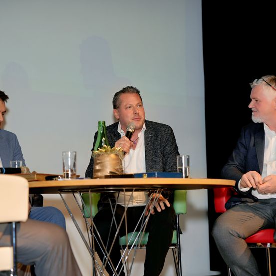 Drei Männer sitzen an einem runden Tisch in einem Konferenzraum. Einer hält ein Mikrofon, ein anderer hat eine grüne Flasche und der dritte lächelt in die Kamera.