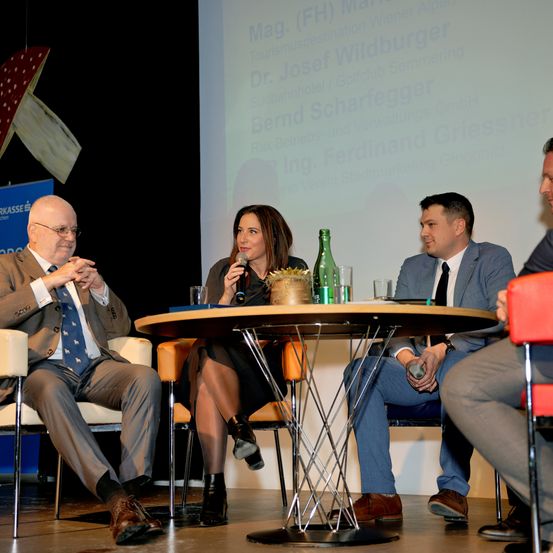 Eine Podiumsdiskussion mit vier Personen, eine hält ein Mikrofon. Ein Tisch mit Gläsern und einer Flasche steht vor ihnen. Eine Projektionsleinwand dahinter mit Text über eine Tour.