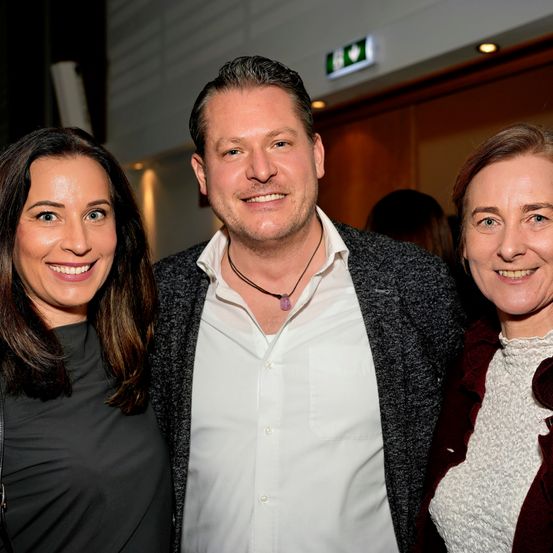 Drei Menschen lächeln für ein Foto; zwei Frauen und ein Mann, möglicherweise bei einer Veranstaltung.
