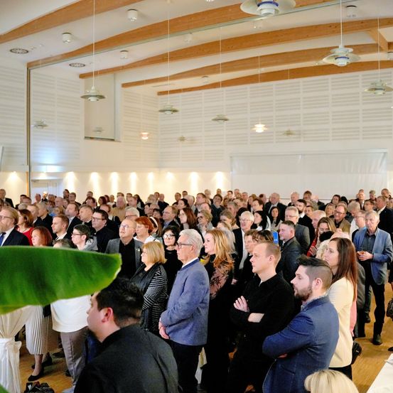Eine große Gruppe von Menschen steht in einem Saal, gekleidet in formelle Kleidung, einige mit Brillen. Der Saal hat Holzbalken und Deckenleuchten. Links sind Tische mit weißen Tischdecken.