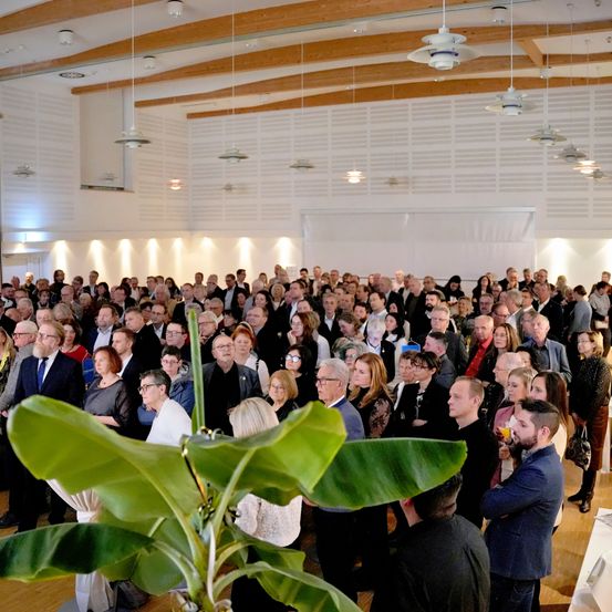 Eine große Gruppe von Menschen ist drinnen versammelt, möglicherweise für eine Veranstaltung. Der Raum hat eine hohe Decke mit Holzbalken und Pendelleuchten. Einige Leute stehen, andere sitzen, und einige tragen Brillen. Eine grüne Pflanze mit großen Blättern ist im Vordergrund.