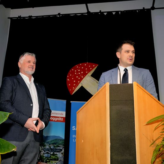 Zwei Männer stehen neben einem Podium; einer spricht in ein Mikrofon und der andere hört zu. Hinter ihnen ist ein Hintergrund mit einem Pilzlogo und einem Banner.