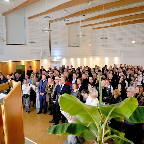 Eine große Gruppe von Menschen steht in einem Saal mit Holzbalken und einem Holzboden. Einige tragen formelle Kleidung, während andere Freizeitkleidung tragen. Eine Frau in einem weißen Shirt steht in der Nähe eines Podiums.