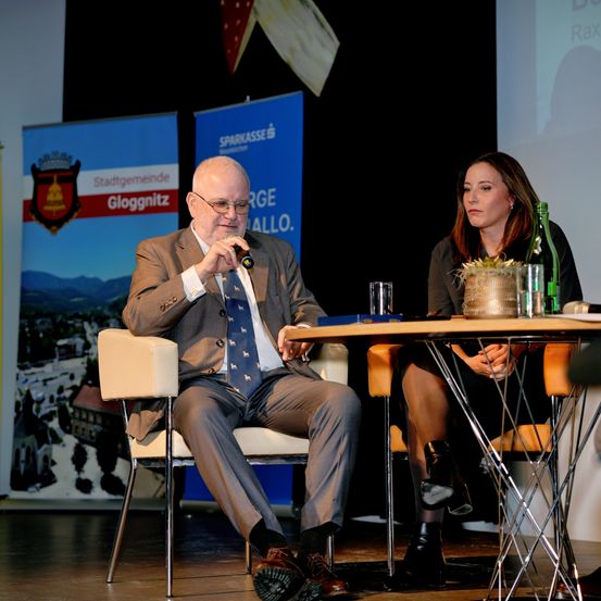 Ein Mann in Anzug spricht in ein Mikrofon an einem Tisch, mit einer Frau, die neben ihm auf einem Stuhl sitzt. Hinter ihnen steht ein Banner mit dem Text Stadtgemeinde Gloggnitz.