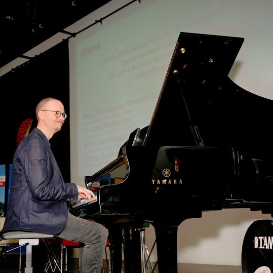 Ein Mann mit Brille und dunklem Anzug spielt ein Yamaha-Klavier auf der Bühne. Dahinter steht eine große Leinwand mit einer Präsentation.