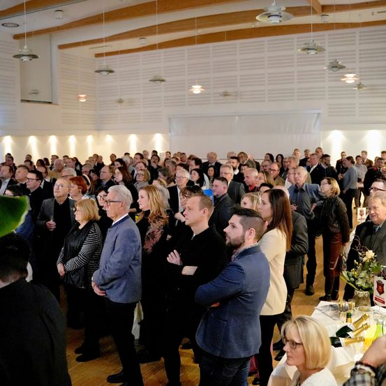 Eine große Gruppe von Menschen steht drinnen, einige schauen auf einen Tisch mit einer Blumendekoration. Der Raum ist gut beleuchtet mit Holzbalken und weißen Wänden.