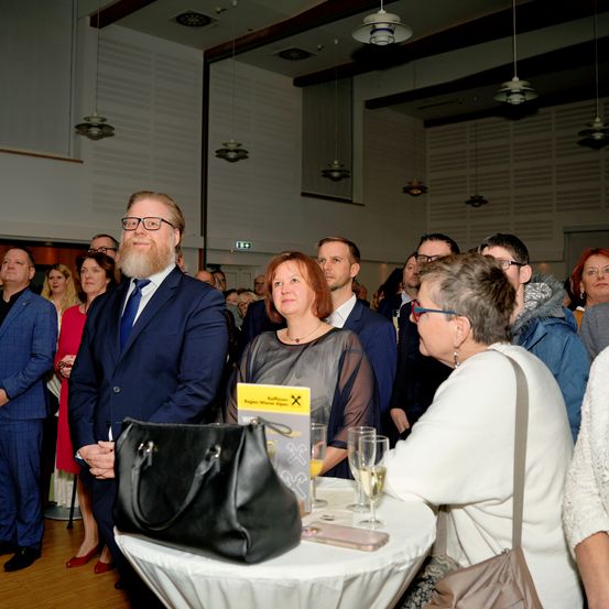 Eine Gruppe von Menschen versammelt sich in einem Raum, möglicherweise bei einer Veranstaltung. Ein Mann mit Bart und Brille steht davor, mit einer Frau in der Nähe. Es gibt Gläser, eine Handtasche und ein Buch auf dem Tisch.