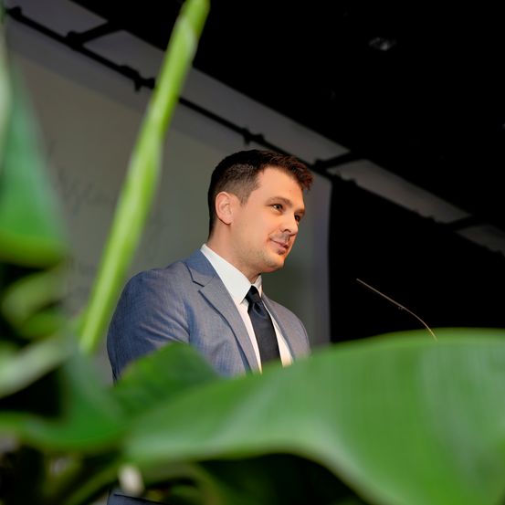 Ein Mann in einem Anzug steht an einem Rednerpult, lächelt und hat ein Mikrofon vor sich. Er ist von großen grünen Blättern umgeben.