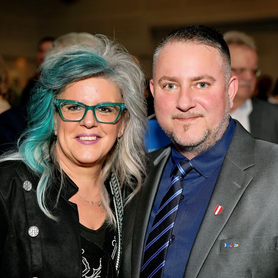 Ein Mann und eine Frau stehen lächelnd zusammen und tragen beide formelle Kleidung. Die Frau hat blaue Haare und eine Brille, und der Mann hat einen Bart und einen Anzug.