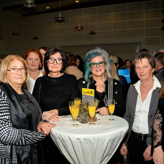 Fünf Frauen stehen lächelnd mit orangefarbenen Getränken an einem runden Tisch. Die Frau auf der linken Seite trägt eine schwarz-weiße Jacke. Die Frau in der Mitte hat blaue Haare. Die Frau auf der rechten Seite trägt eine graue Jacke. Hinter ihnen stehen und sprechen weitere Personen.
