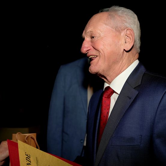 Ein älterer Mann in einem dunkelblauen Anzug und roter Krawatte, lächelt und hält ein Stück Papier in der Hand, möglicherweise bei einer Veranstaltung.