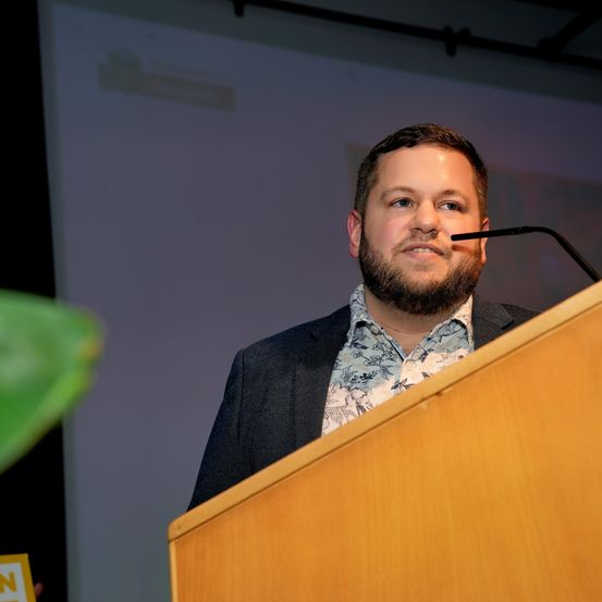 Ein Mann mit Bart steht an einem Podium und spricht in ein Mikrofon in einem Konferenzraum.