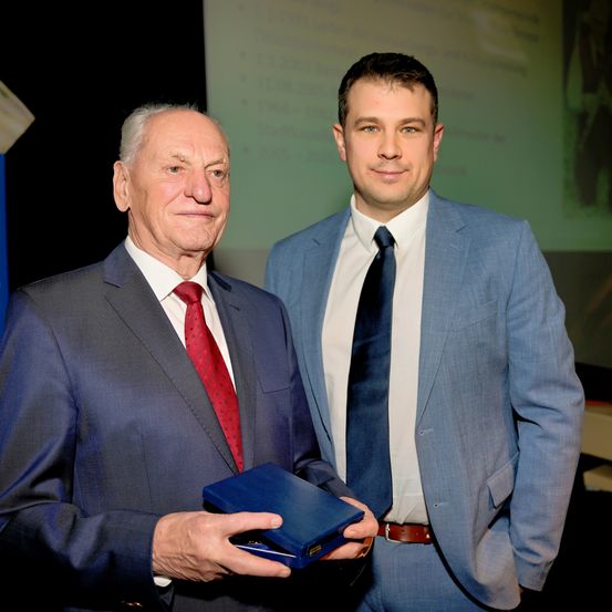 Zwei Männer in Anzügen und Krawatten, einer älter und einer jünger, stehen nebeneinander und halten eine blaue Mappe. Der ältere Mann lächelt, während der jüngere Mann in die Kamera schaut. Hinter ihnen befindet sich eine Projektionsleinwand mit grünem Hintergrund.