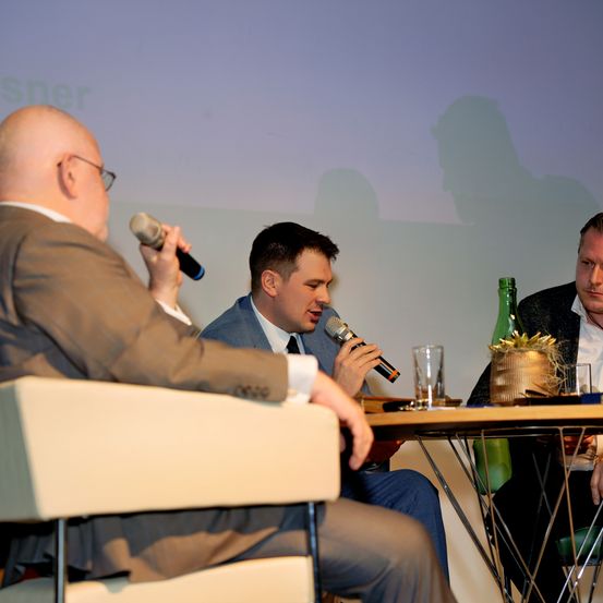 Drei Männer sitzen an einem Tisch, einer hält ein Mikrofon, während ein anderer spricht. Der Raum ist durch eine Projektionsleinwand im Hintergrund beleuchtet.