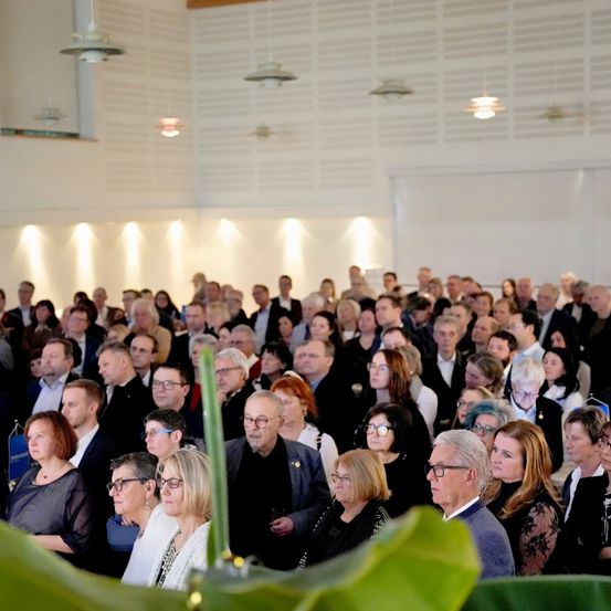 Eine große Gruppe von Menschen ist drinnen versammelt, wahrscheinlich in einem Konferenzsaal. Sie sind formell gekleidet, viele tragen Brillen. Ein grüner Pflanzen ist im Vordergrund sichtbar.