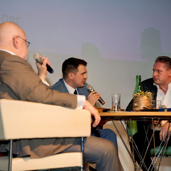 Drei Männer in Anzügen sitzen mit Mikrofonen an einem Tisch, möglicherweise für ein Interview. Der Mann auf der linken Seite trägt eine Brille und hält ein Mikrofon. Vor ihnen steht ein Glas und eine Flasche. Hinter ihnen zeigt eine Projektionsleinwand Schatten.